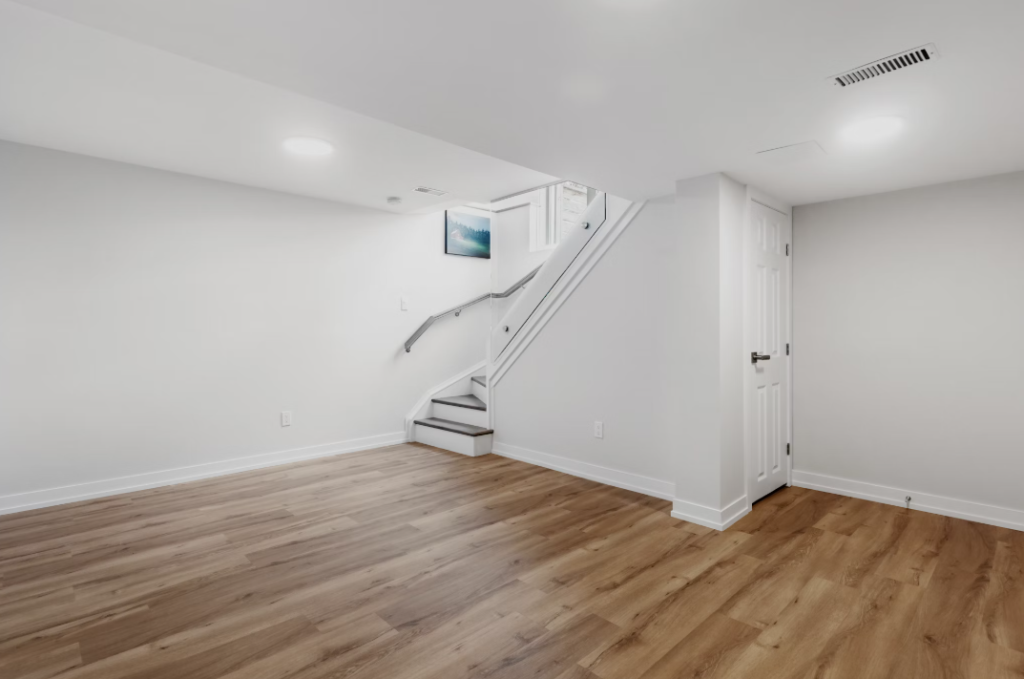 An empty basement with light wood flooring, white walls, and a staircase.