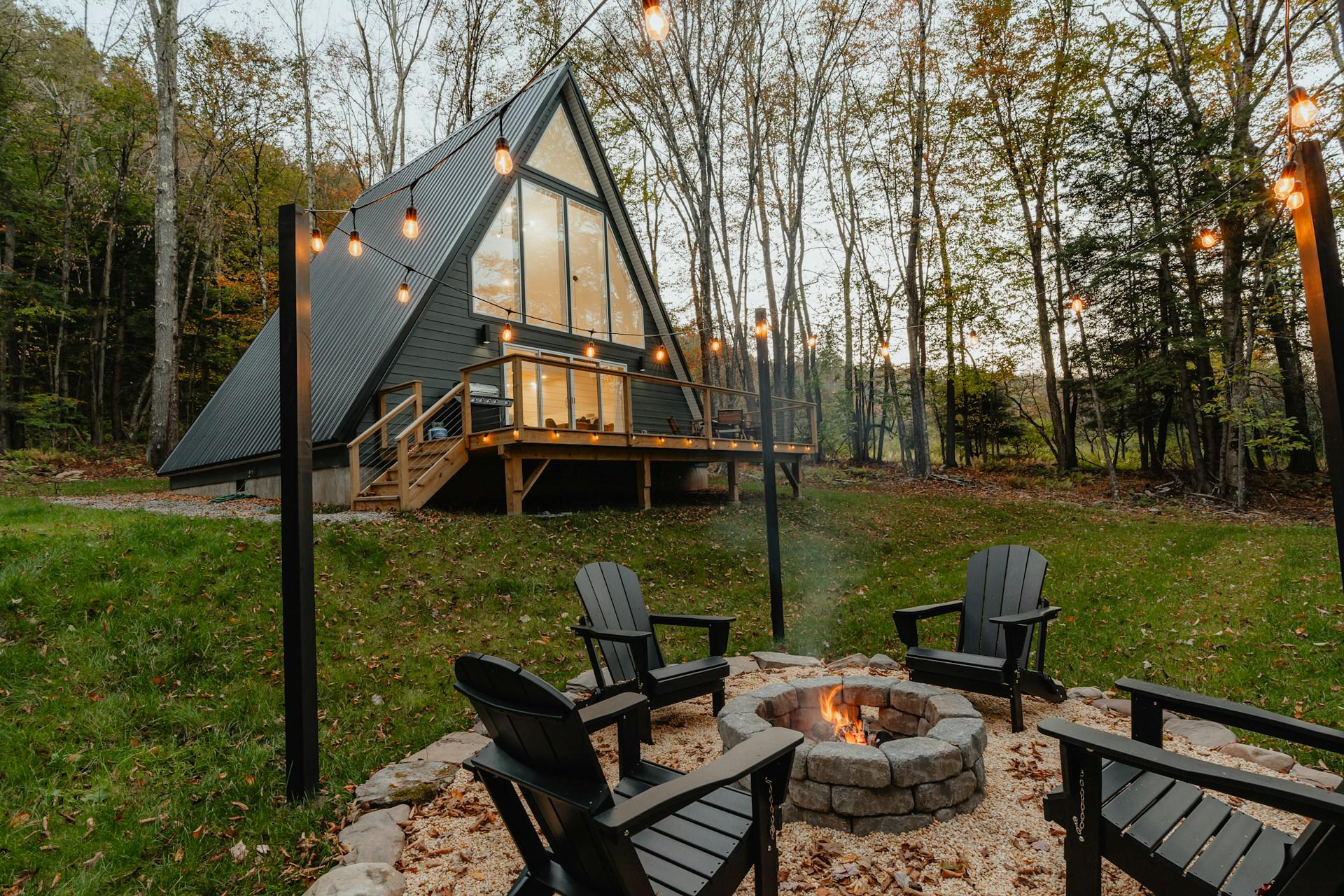 A triangle-shaped cabin can be seen with a sitting area outside it with fairy lights