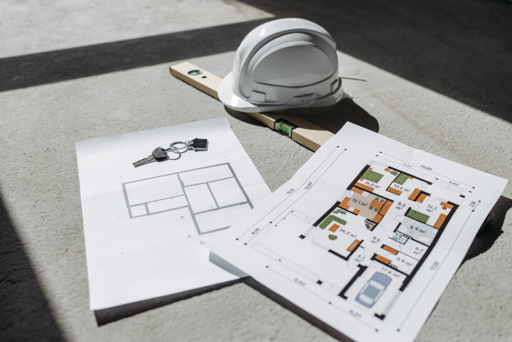 A construction helmet, a leveling tool, house keys, and floor plans lying on a concrete surface, representing the planning stage of a custom home build.