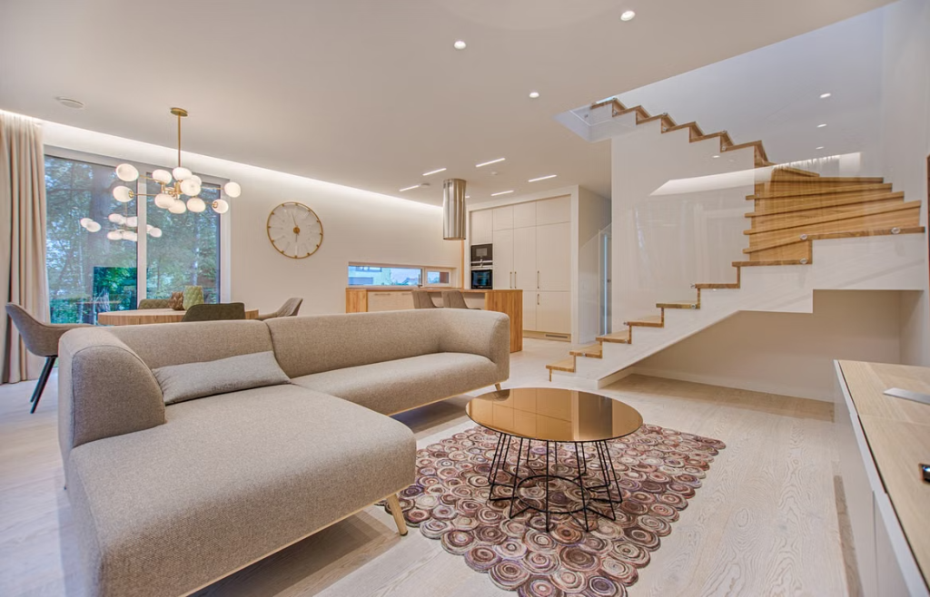 Spacious modern open-plan living room with a beige sectional sofa, glass-railing staircase, round coffee table, and kitchen area in the background